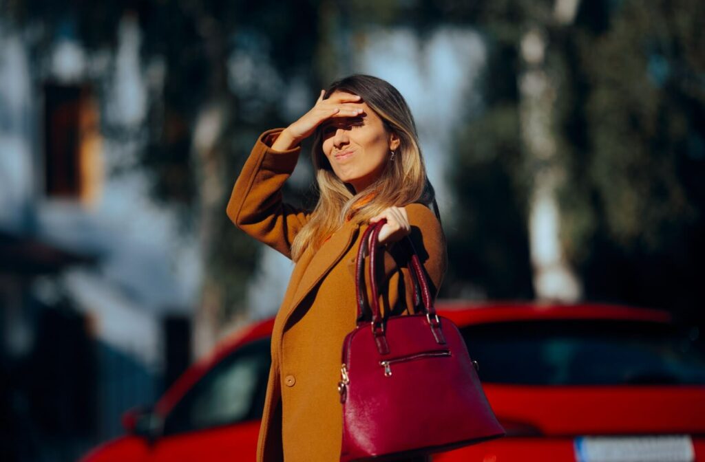 A woman standing outdoors squinting and shielding her eyes from bright sunlight with her hand.