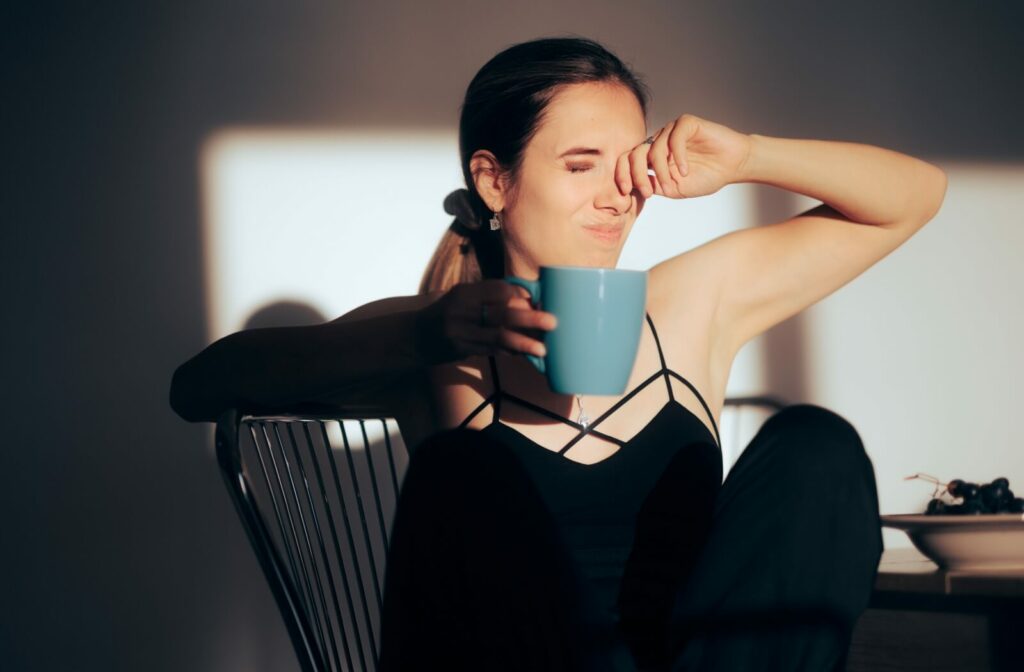 A woman sitting at a table holding a mug and rubbing her eyes as bright indoor light creates harsh shadows behind her.